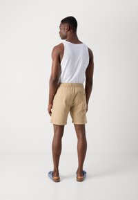 Beige badebukser med elastisk talje, baglomme, båret med en hvid tanktop og blå slip-on sko, set bagfra.