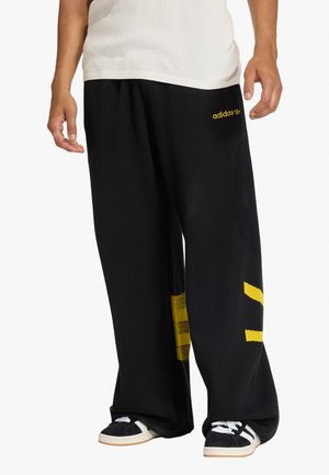 Persona che indossa pantaloni neri Adidas a gamba larga con strisce gialle e logo, scarpe da ginnastica bianche con strisce nere e un top bianco.