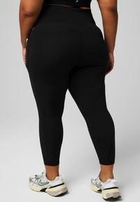 Schwarze Sportleggings mit hohem Bund, glatter Textur und figurbetontem Schnitt, kombiniert mit weißen und silbernen Sportschuhen.
