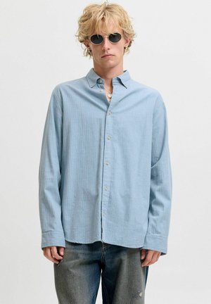 Personne blonde portant des lunettes de soleil rondes noires, une chemise bleu clair à boutons et un jean bleu délavé, debout devant un fond clair uni.