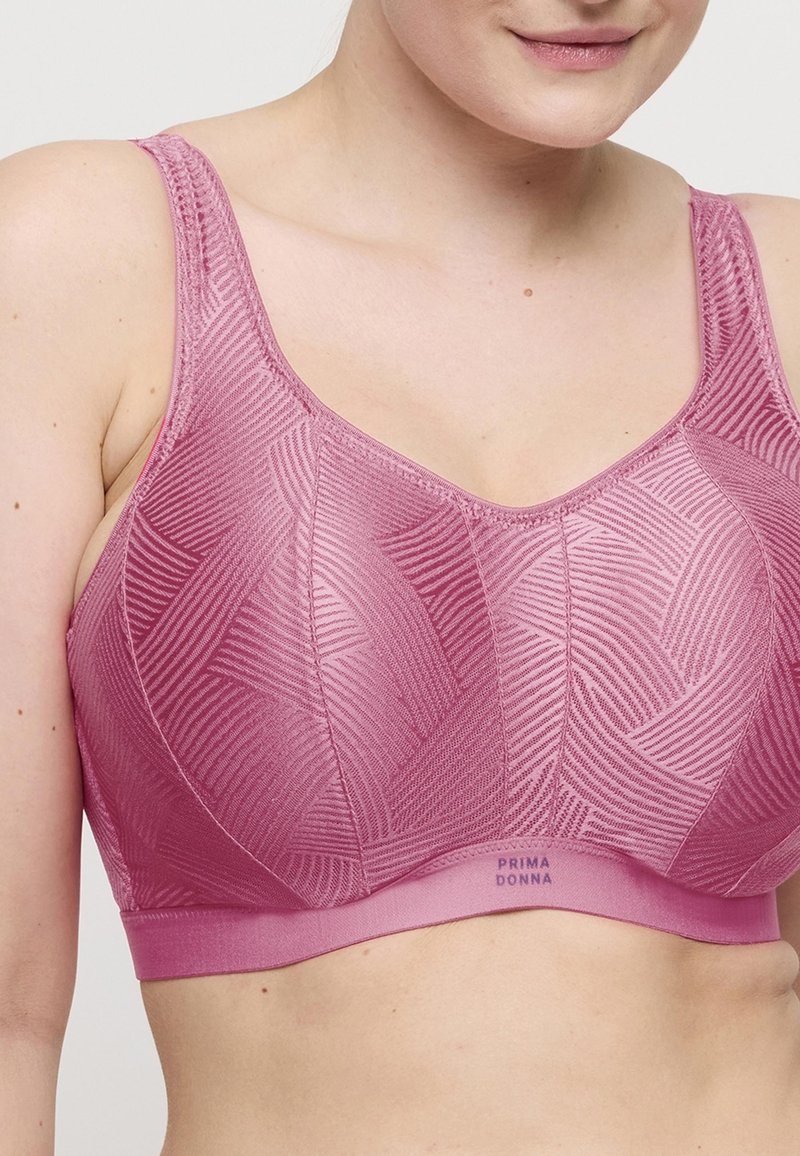 Nainen, joka on pukeutunut vaaleanpunaisiin kuviollisiin pehmeisiin liiveihin, joissa on leveät hihnat, ilman rautalankatukea, ja "Prima Donna" -brändätty elastinen nauha kuppeja alla.
