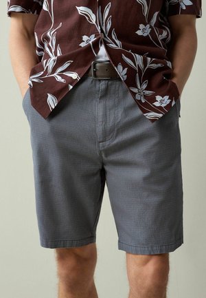 Mann trägt graue, strukturierte Shorts und ein braunes, geblümtes Hemd mit Knöpfen, das nicht in die Hose gesteckt ist, die Hände in den Taschen, steht vor einem neutralen Hintergrund.
