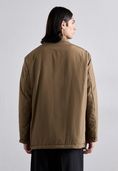 Chaqueta marrón acolchada con corte recto, que presenta un cuello alto y mangas largas. Textura suave, diseño minimalista y sin herrajes visibles.
