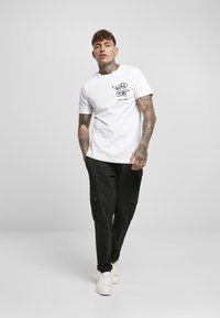 White cotton t-shirt with black graphic print on the chest. Slim fit, crew neck design. Paired with black cargo pants and white sneakers.