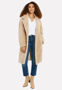 Manteau teddy en peluche beige avec un col oversized, associé à une chemise blanche à col en V et un jean bleu. Le modèle se tient dans des bottines beige à la cheville.