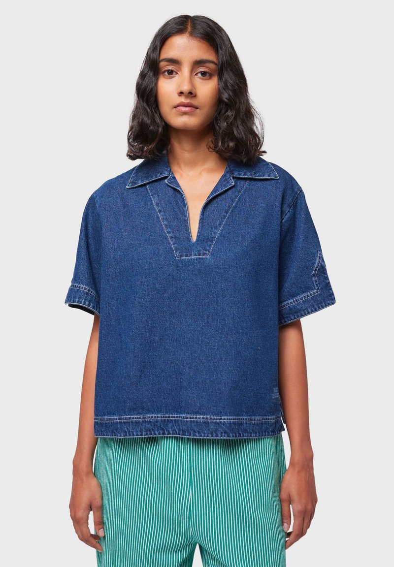 Camicia in denim a maniche corte con scollo a V, caratterizzata da una vestibilità rilassata e tasche applicate. Il design è di colore blu scuro con cuciture a contrasto.
