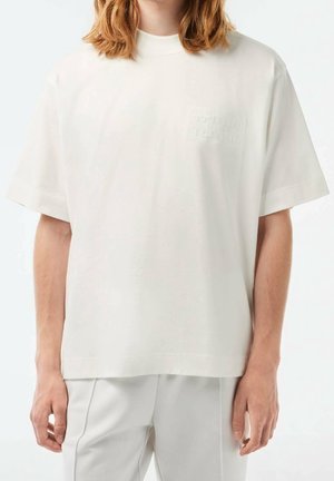 T-shirt en coton blanc avec un col montant et des manches courtes ; présente un logo discret embossé sur la poitrine. Texture douce, coupe décontractée.