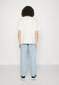 Camiseta de algodón beige claro, de corte relajado, con mangas cortas. Combinada con jeans rectos de un azul claro y zapatillas deportivas negras.