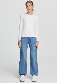 Biała koszula z długim rękawem zestawiona z luźnymi niebieskimi spodniami cargo z dużymi bocznymi kieszeniami, dopełniona białymi trampkami.