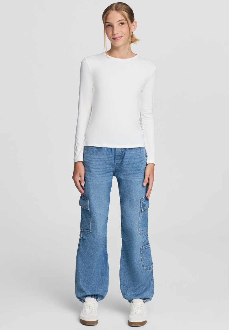 Biała koszula z długim rękawem zestawiona z luźnymi niebieskimi spodniami cargo z dużymi bocznymi kieszeniami, dopełniona białymi trampkami.