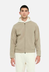 Beige zip-up hoodie med struktureret materiale, hvid hætteforing og elastiske manchetter. Modellen har cremede bukser på. Minimalistisk design.