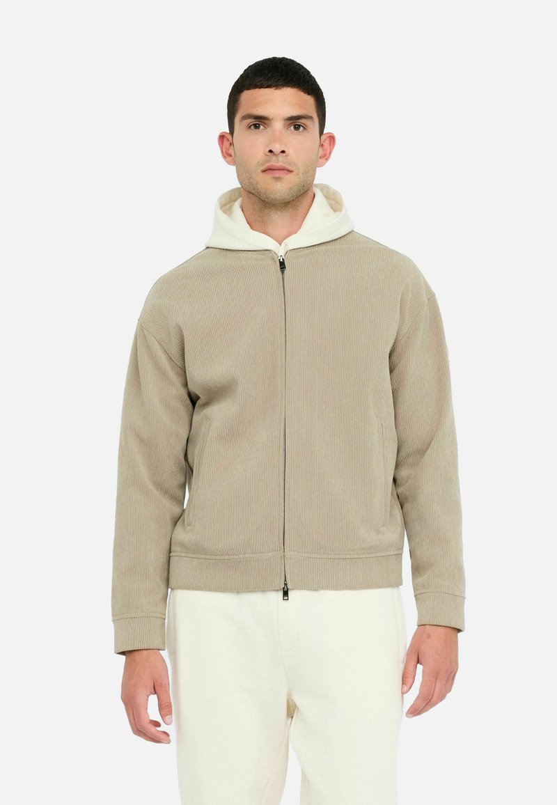 Beige zip-up hoodie med struktureret materiale, hvid hætteforing og elastiske manchetter. Modellen har cremede bukser på. Minimalistisk design.