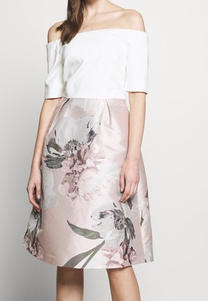 Vrouw draagt een witte off-shoulder top en een knielange roze rok met een grote bleke bloemenprint tegen een effen achtergrond.
