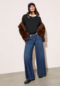 Crna majica s bijelim ovratnikom, smeđi umjetni krzneni jakna, široke plave traperice i crni loafers. Model nosi plavu kapu.