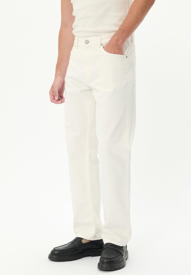 Pantalones de denim blanco con un diseño de pierna recta, que cuentan con cinco bolsillos y un cierre de botón. Llevados con zapatos negros sin cordones.