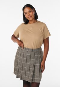 Beige T-Shirt mit kurzen Ärmeln und dezentem Logo, kombiniert mit einem grauen, karierten, gefalteten Rock mit hoher Taille und schwarzen Musterakzenten.