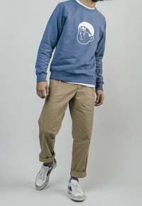 Modrá mikina s bílým potiskem obličeje, spojená s béžovými chinos kalhotami s rolled-up stylem a bílými teniskami s navy akcenty.