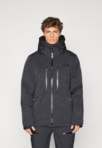 Giacca nera impermeabile con colletto alto, cappuccio regolabile e due tasche sul petto. Caratterizzata da chiusure con zip e dettagli con logo.