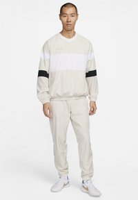 Completo da allenamento Nike beige con una striscia orizzontale bianca e dettagli neri. Presenta polsini e vita elasticizzati. Indossato con scarpe da ginnastica bianche.