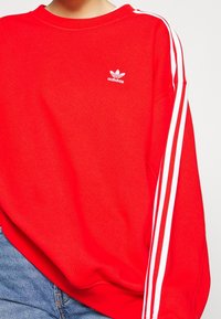 Sweatshirt rouge Adidas avec un design à trois bandes blanches sur les manches et un petit logo Adidas blanc sur la poitrine, associé à un jean bleu.