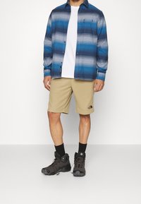Man som bär en blårandig skjorta med knappar, vit T-shirt, beige knävärmande shorts, svarta strumpor och svarta vandringsskor, stående mot vit bakgrund.