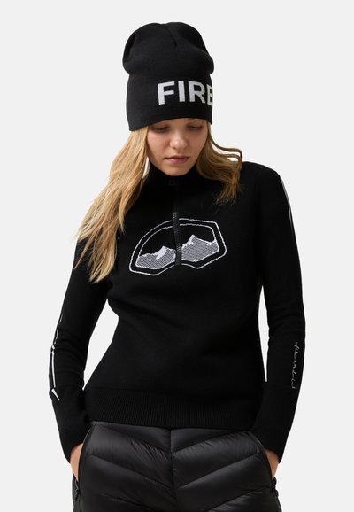 Fekete kötött pulóver magas gallérral, fehér grafikai mintával és hímzett részletekkel az ujjain. Fekete puffadt nadrággal párosítva.