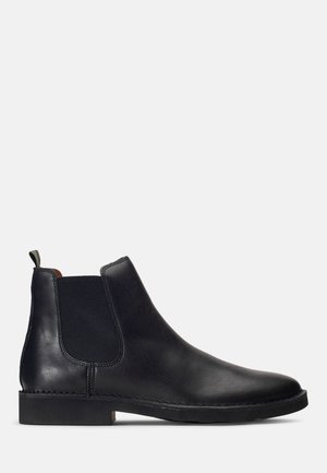 Botas Chelsea pretas em couro com painéis elásticos laterais, textura suave, bico arredondado e sola de borracha plana, apresentando uma pequena aba para puxar.