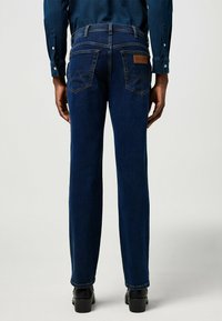 Dark blue denim jeans with a straight cut, featuring angled back pockets and a leather logo patch on the waistband. Smooth texture.