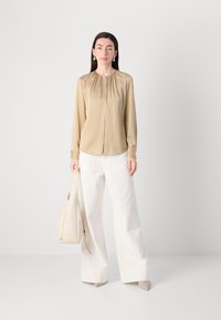 Blusa di raso beige con scollatura arricciata, maniche lunghe e chiusura a bottoni, abbinata a pantaloni bianchi a gamba larga e una borsa beige.