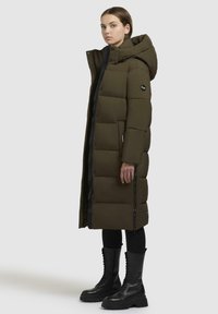 Long olive green puffer coat with a hood, featuring a zip closure, side pockets, and a quilted design. Worn with black lace-up combat boots.
