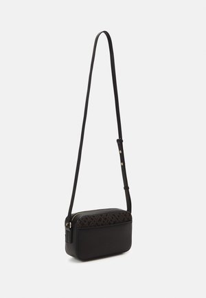 Lille sort crossbody taske med justerbar rem, vævet tekstureret topsektion og lynlåslukning mod en ensfarvet lys baggrund.
