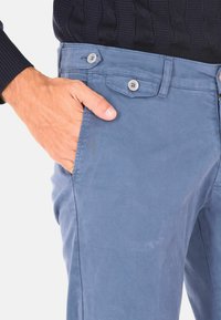 Pantaloni di cotone azzurri chiari con una superficie liscia, dotati di due bottoni in vita e di una tasca laterale, mostrati con una mano in tasca.