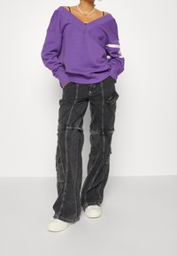 Lila oversized sweatshirt med djup V-hals och vita ränder på ärmarna, tillsammans med svarta cargo-byxor med flera fickor och kontrasterande sömmar.