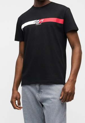 Mand iført en sort Tommy Jeans t-shirt med hvide og røde horisontale striber over brystet, kombineret med lyseblå jeans.