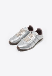 Srebrne metaliczne sneakersy o gładkim wykończeniu, beżowe sznurówki i brązowa gumowa podeszwa. Posiadają przeszywane akcenty i okrągły kształt noska.