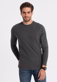 Dlouhá t-shirt s kulatým výstřihem v tmavě šedé barvě, vyrobená z hladkého materiálu. Nošena s volnými modrými džíny. Žádné viditelné vzory nebo akcenty.
