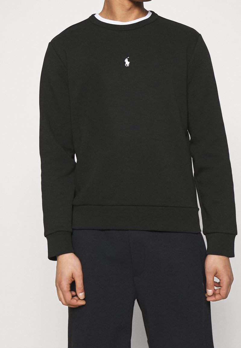 Homme portant un sweat-shirt noir à manches longues et col rond avec un petit logo blanc de joueur de polo sur la poitrine.