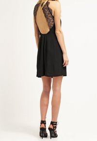 Robe noire avec dos en dentelle, design sans manches et jupe évasée. Portée avec des chaussures à talons hauts, mettant en valeur les jambes.