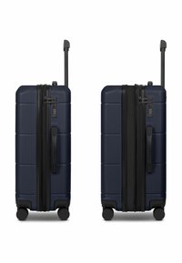 Maleta rígida azul oscuro con superficie texturizada, ruedas dobles, asa telescópica y cerradura de combinación. La cremallera corre verticalmente a lo largo del costado.