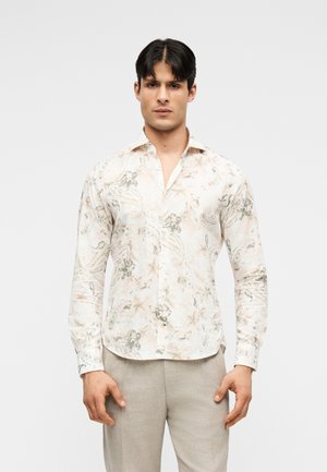 Άνδρας που φοράει μπεζ πουκάμισο με μακρύ μανίκι και λουλουδάτο σχέδιο και ανοιχτό γκρι παντελόνι, στέκεται μπροστά σε απλό ανοιχτό γκρι φόντο.