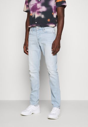 Muž nosící úzké džíny světle modré barvy, bílé tenisky a košili s krátkým rukávem a abstraktními fialovo-oranžovými vzory.