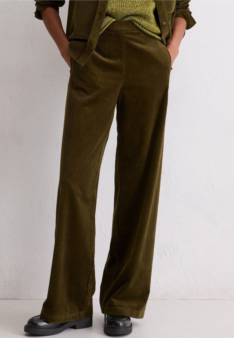 Personne portant un pantalon en velours côtelé vert olive à jambes larges, un pull en maille assorti et des chaussures noires épaisses, debout les mains dans les poches.