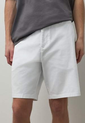 Mand iført almindelige hvide knælange shorts og en mørkegrå t-shirt, stående med armene ned langs siderne.