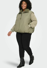 Smilende kvinne som går, iført lys grønn vattert vest med leopardmønstret krage, lys grønn genser, svarte leggings og svarte ankelstøvler.