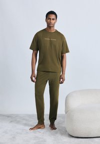 Σετ απόλαυσης ελιάς πράσινο t-shirt και παντελόνι από μαλακό ύφασμα, με μικρό λογότυπο στο πουκάμισο. Το μοντέλο στέκεται ξυπόλυτο σε ένα ανοιχτό χαλί.