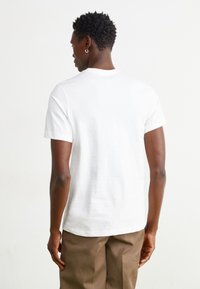 Camiseta blanca de manga corta con cuello redondo, hecha de una tela suave y ligera, combinada con pantalones beiges, que muestra un diseño sencillo.