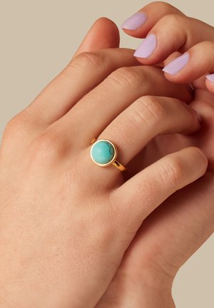Main aux ongles peints en lavande portant une bague en or ornée d'une pierre turquoise ronde à l'annulaire, sur fond beige.