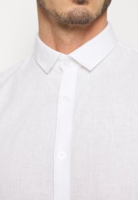 Camicia bianca di lino con colletto classico, chiusura a bottoni e finitura testurizzata. Il tessuto appare leggero e traspirante.