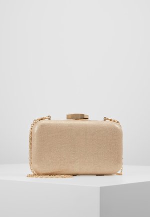 Pochette rectangulaire à paillettes dorées avec bords arrondis et une bandoulière chaîne dorée, posée sur un piédestal blanc devant un fond uni.