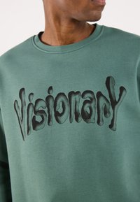 Felpa verde con scollatura rotonda, caratterizzata da un testo ondulato nero "Visionary" sul davanti. Realizzata in un morbido tessuto testurizzato.